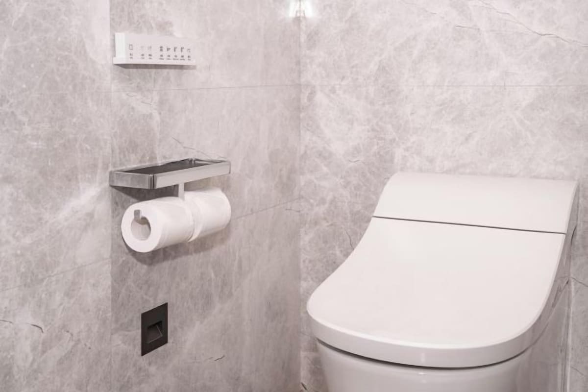 Un baño moderno con inodoro, dispensador de toallas de papel y botón de descarga.
