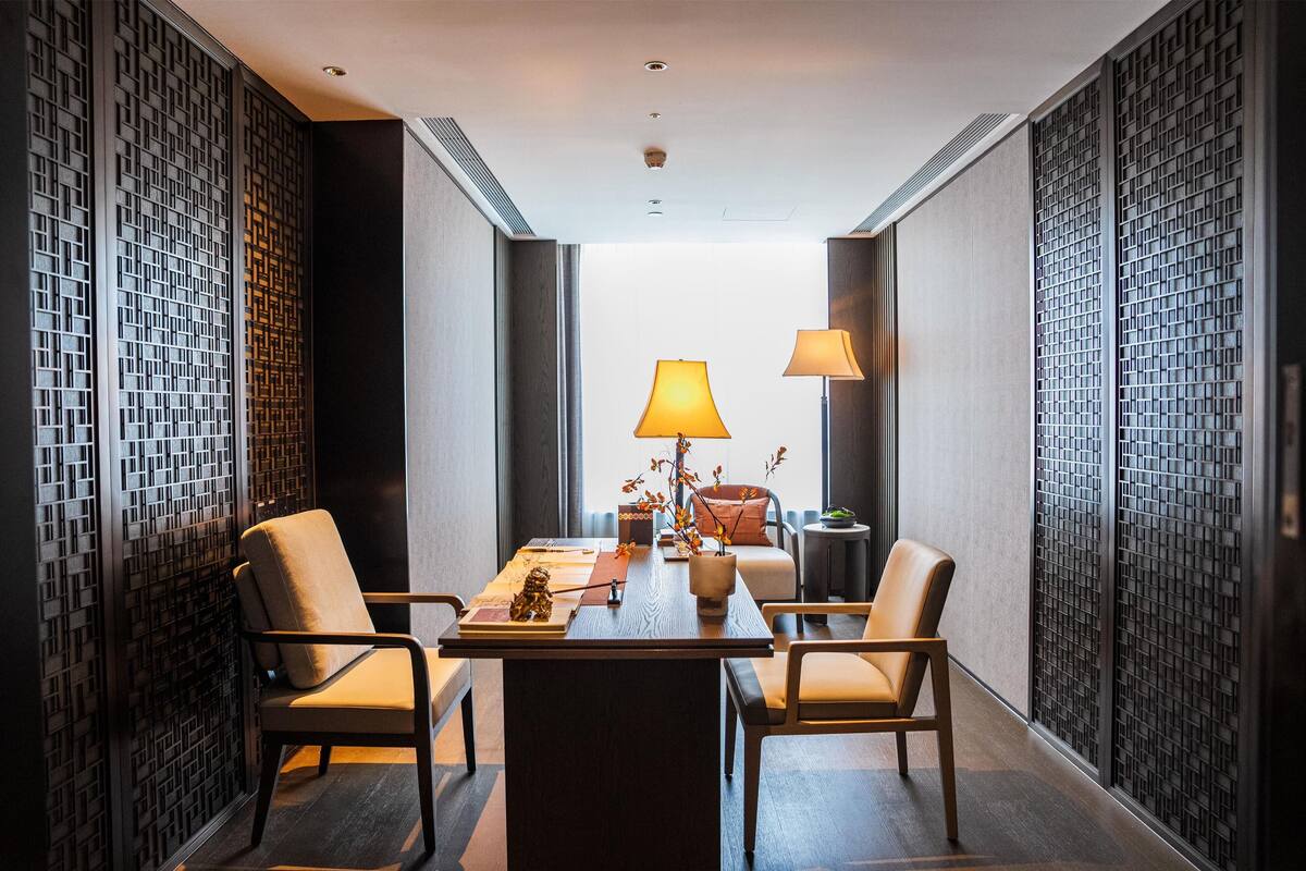 A modern hotel room with a dining area, featuring a wooden table, two chairs, a sofa, and a decorative plant.