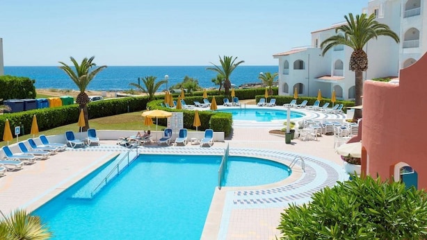 Uma área de piscina com espreguiçadeiras e guarda-sóis, vista desobstruída para o mar e um edifício com arcos.