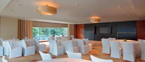 A conference room with round tables and white chairs, a large window with curtains, and a flat-screen TV on the wall.