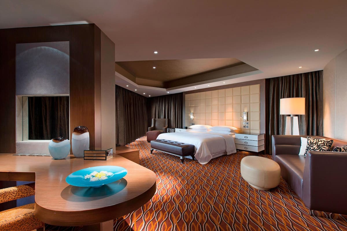 Una habitación de hotel moderna con una cama grande, un área de descanso con una mesa y vistas a la ciudad a través de la ventana.