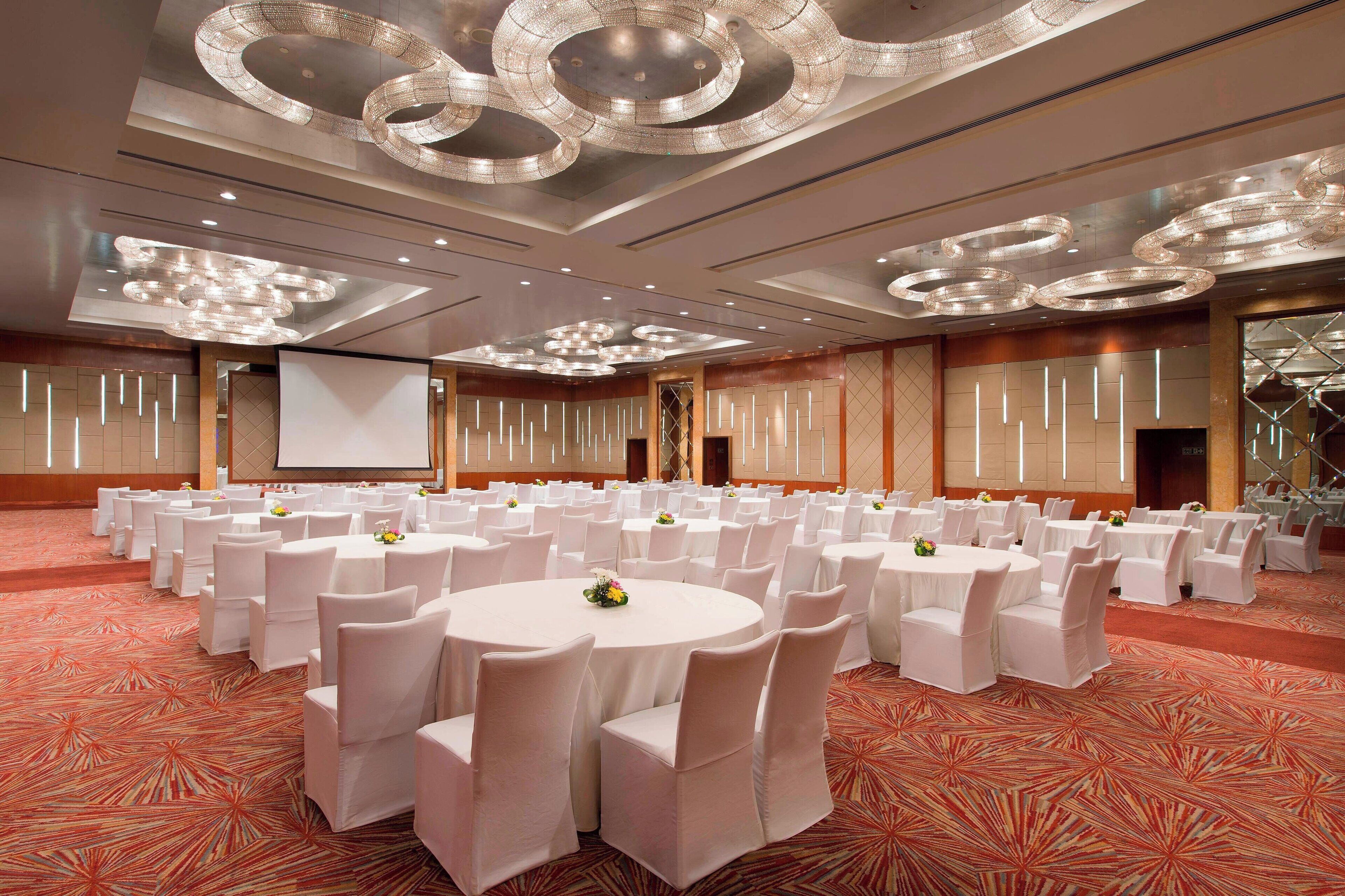 A banquet hall with round tables and white chairs, a large screen, and a patterned carpet.