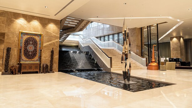 Un vestíbulo moderno con suelo de mármol, un gran tapiz decorativo, una escalera y una fuente de agua.