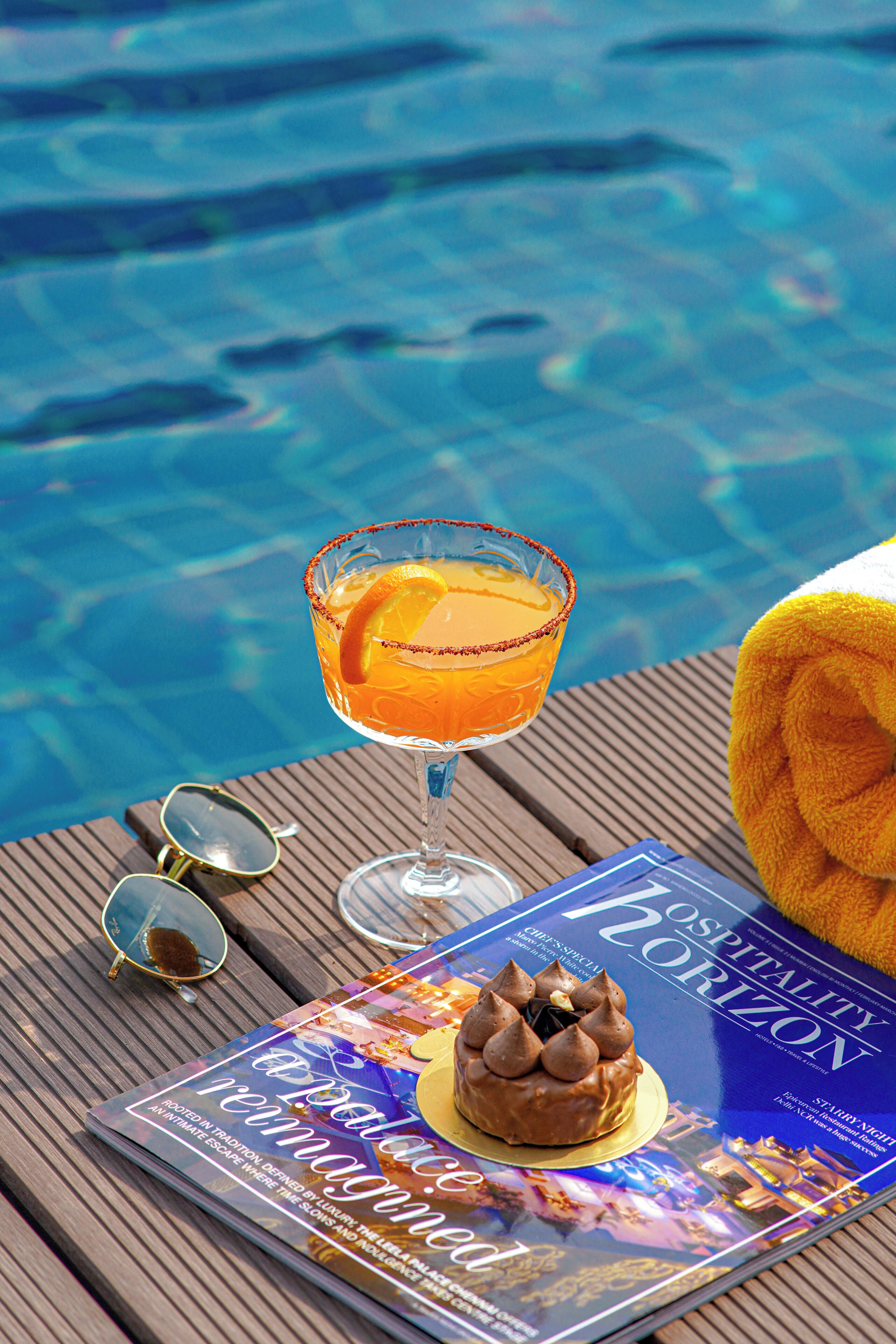 A magazine on a wooden table, a cocktail glass with an orange slice, sunglasses, and a towel.
