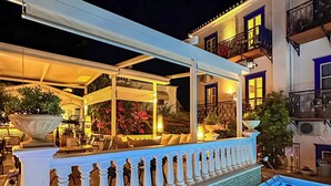 Un espace aménagé au bord de la piscine, avec un coin salon couvert, une balustrade blanche et un bâtiment avec des balcons en arrière-plan.