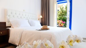 Un lit bien fait, avec une tête de lit blanche et des oreillers, une table de chevet avec une lampe, et une fenêtre donnant sur un paysage verdoyant et fleuri.