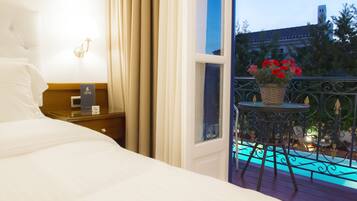 A hotel room with a bed, a nightstand, a lamp, a window with curtains, and a balcony with a table and flowers.