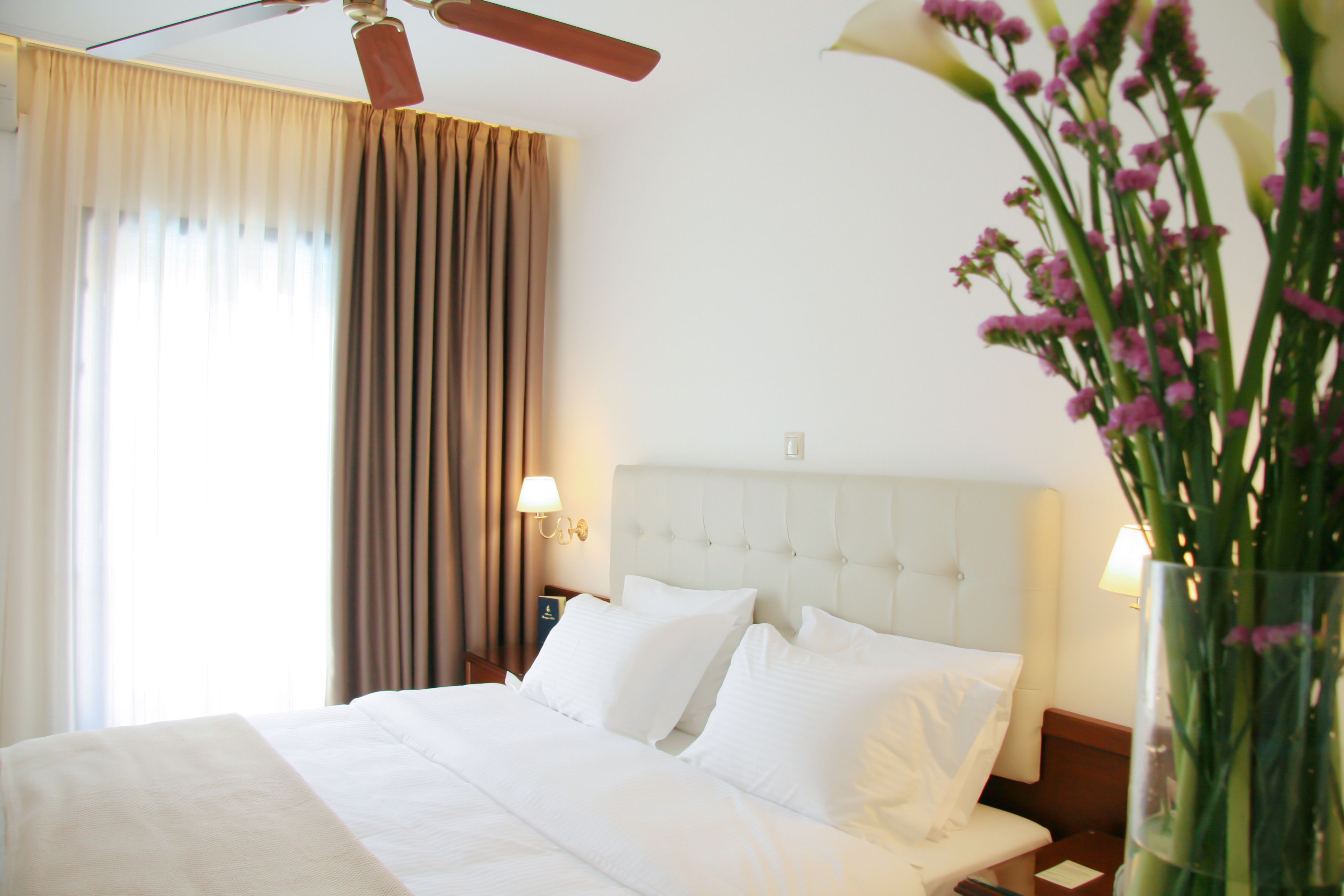 Una habitación de hotel con cama, cortinas, mesita de noche, lámpara y un jarrón con flores.