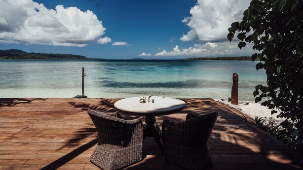 Una terraza de madera con sillas de mimbre y una mesa, con vistas a un cuerpo de agua tranquilo.