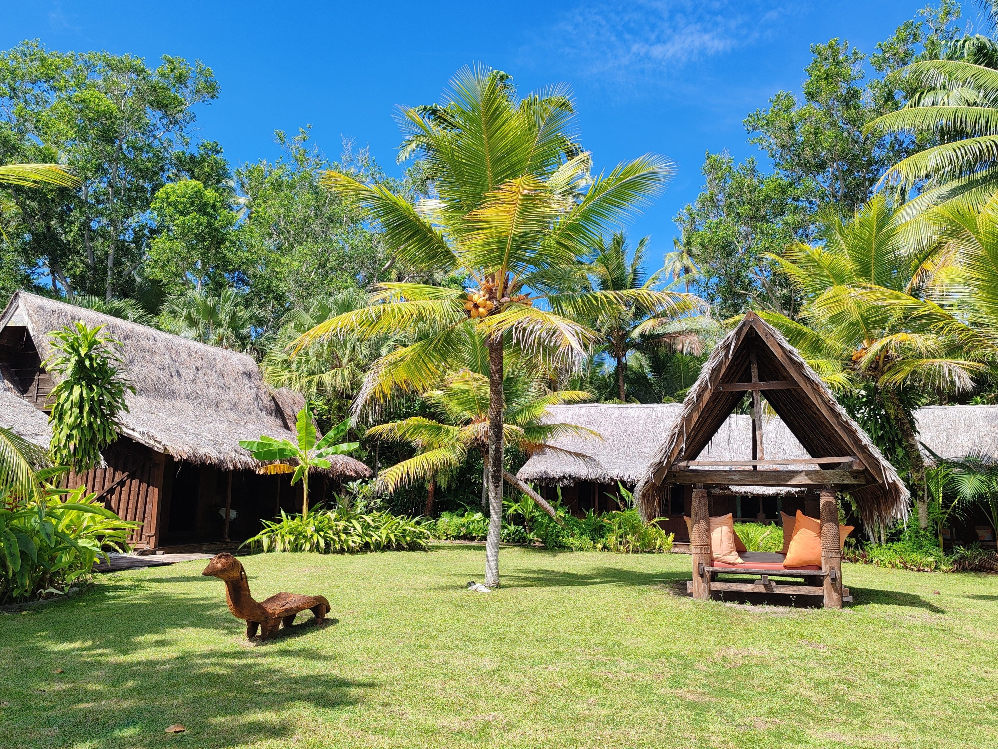 A tropical resort with thatched-roof huts, a wooden lounge chair, and lush greenery.