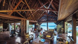 Amplio espacio interior con muebles de madera, un gran ventanal con vista al océano y una estructura de techo de paja.
