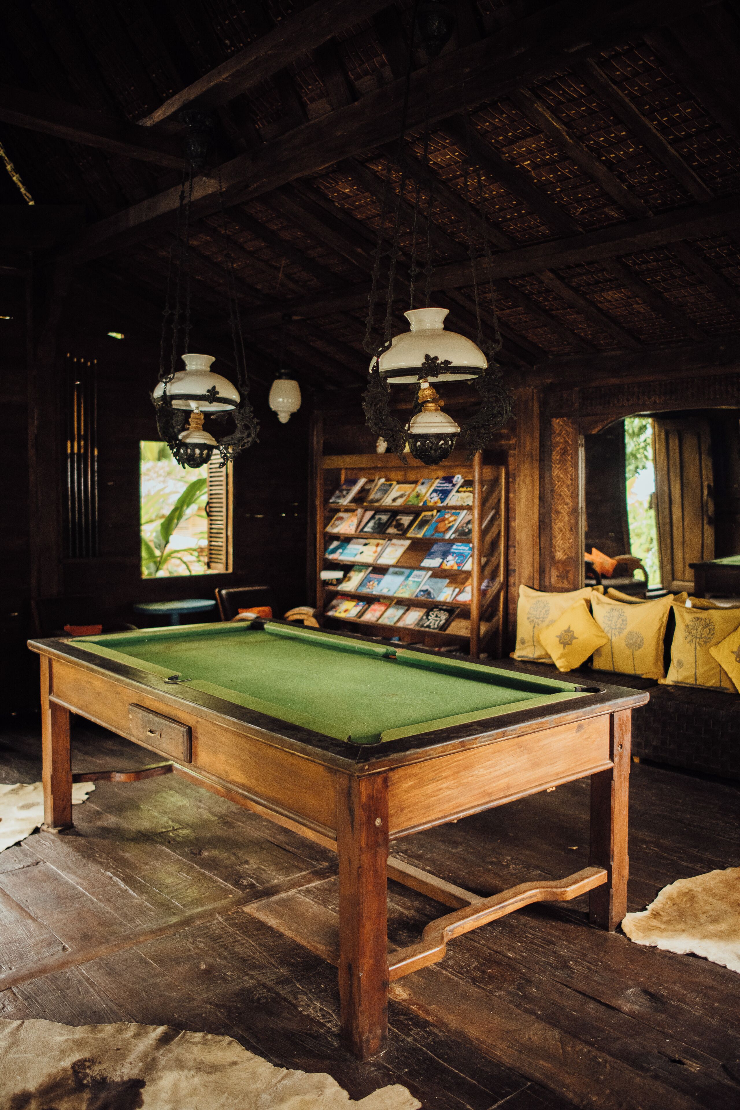 Une table de billard dans une pièce au plancher en bois, une bibliothèque remplie de livres et des lampes suspendues.