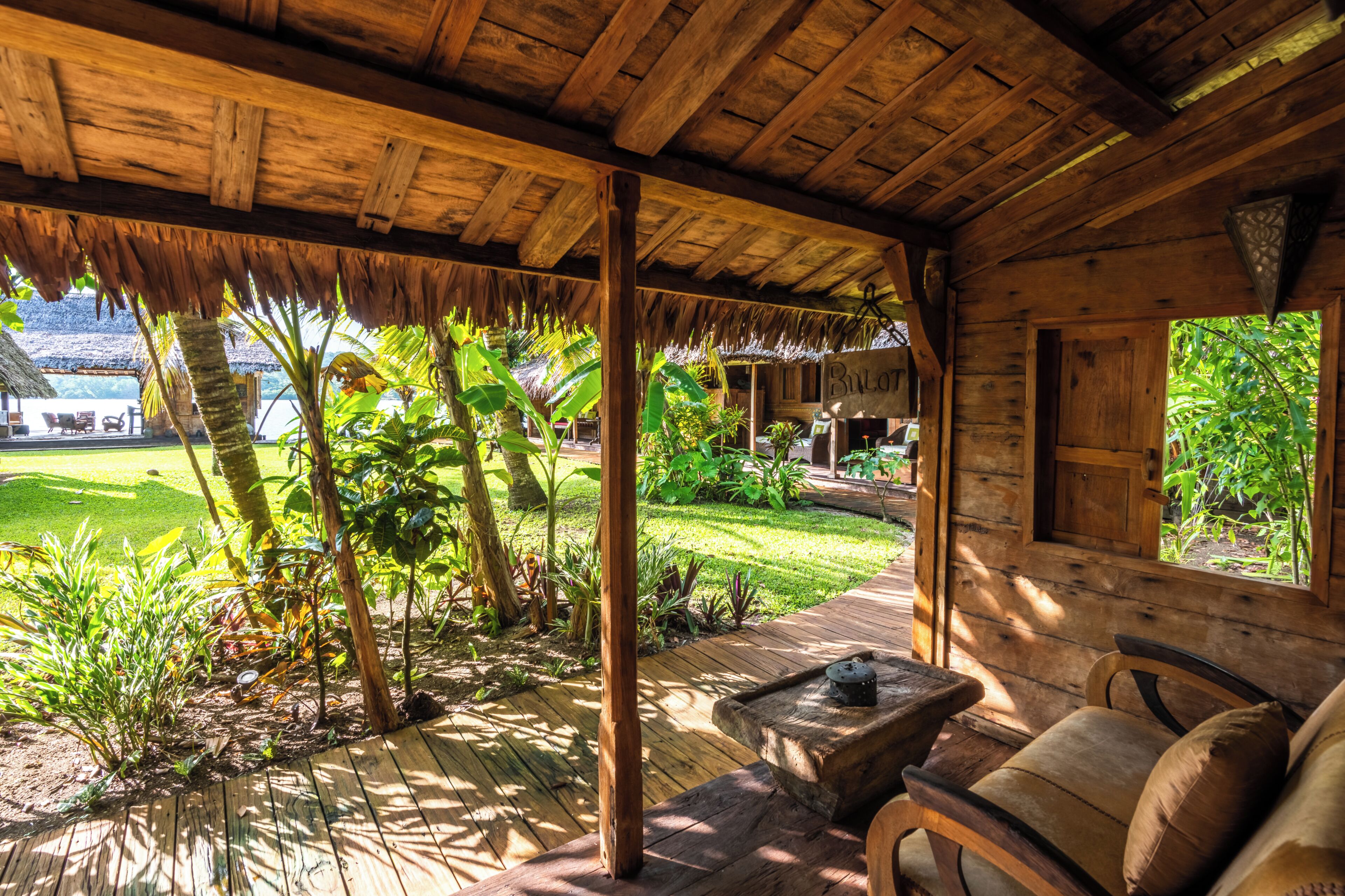 Una cabaña de madera con techo de paja, una terraza de madera y vistas a un jardín exuberante y un edificio a lo lejos.