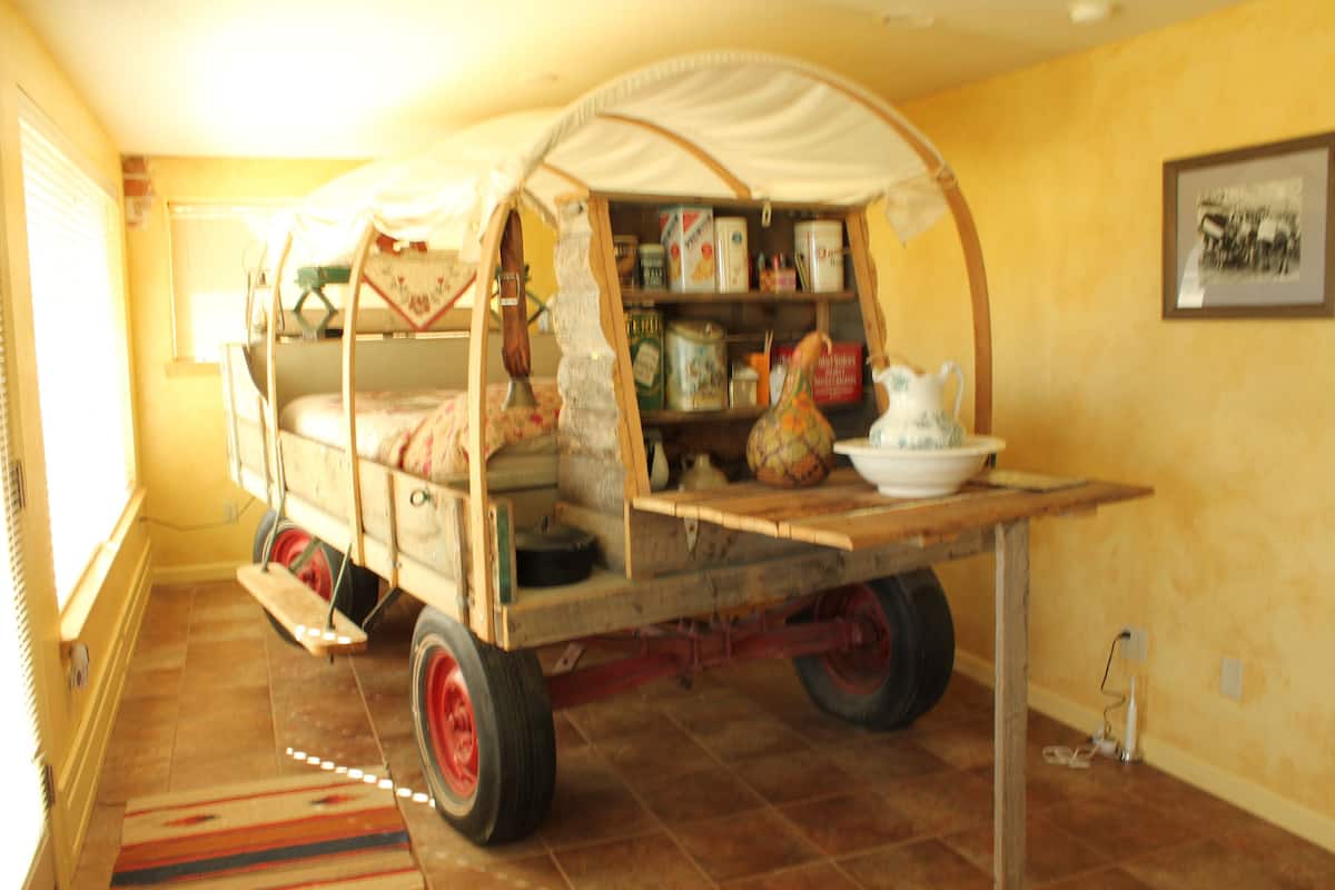 Um carrinho de madeira antigo com toldo, repleto de diversos objetos, posicionado em um cômodo com paredes amarelas e piso de azulejos.