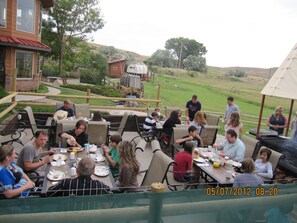An outdoor dining area with multiple tables and chairs, surrounded by a grassy landscape and trees.