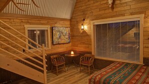 Una habitación acogedora de estilo cabaña con una escalera de madera, chimenea, dos sillones, cama con colcha estampada y un ventanal con persianas.