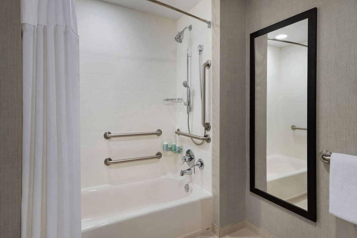 Bathroom features a bathtub, large mirror, and shower area with a tiled floor and handrails for safety.