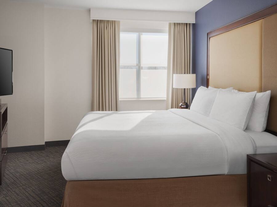 A hotel room with a large bed, a television on a wooden stand, a window with curtains, and a nightstand with a lamp.