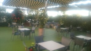 Een terras buiten met tafels en stoelen, omringd door groen en plantenpotten.