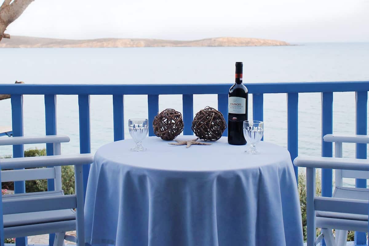 Une table dressée avec une bouteille de vin, deux verres et des objets décoratifs, le tout posé sur une nappe bleue, avec vue sur une étendue d’eau et une terre lointaine.