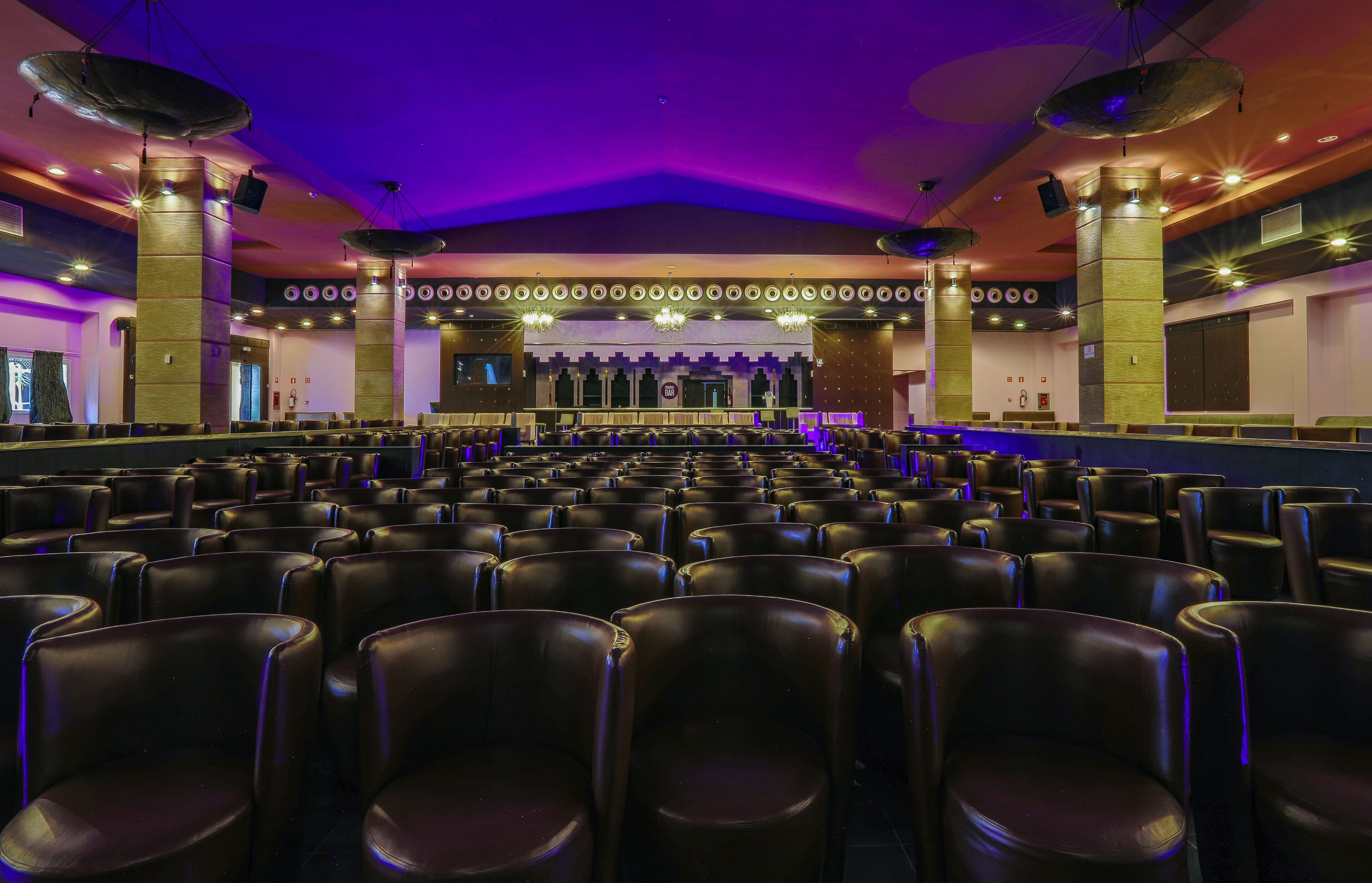 A theater with rows of black leather seats, a stage with a backdrop, and a ceiling with circular lights.