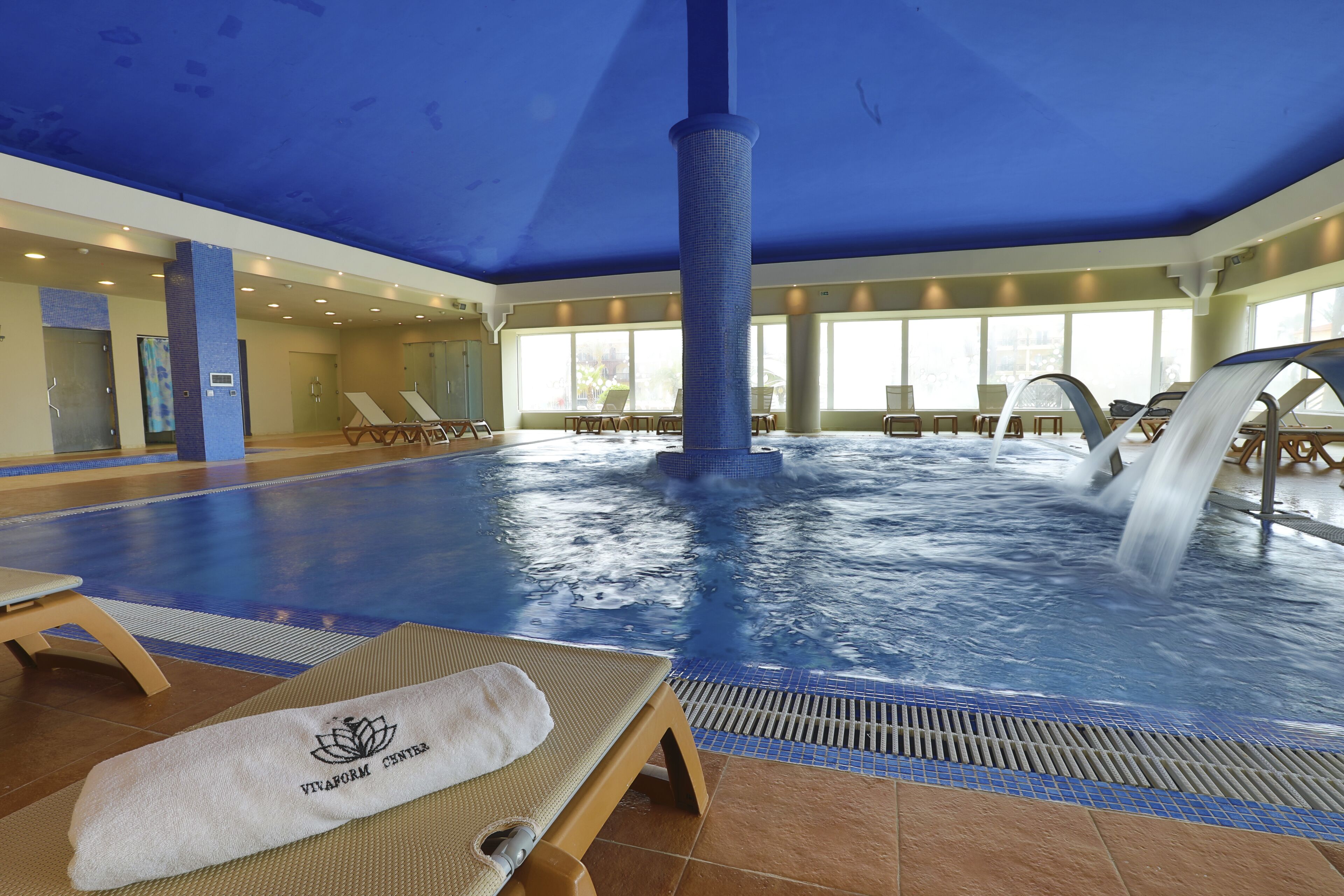 Una amplia piscina interior con cascada, sillas de descanso y una toalla con la marca 'VivaForm Center'.