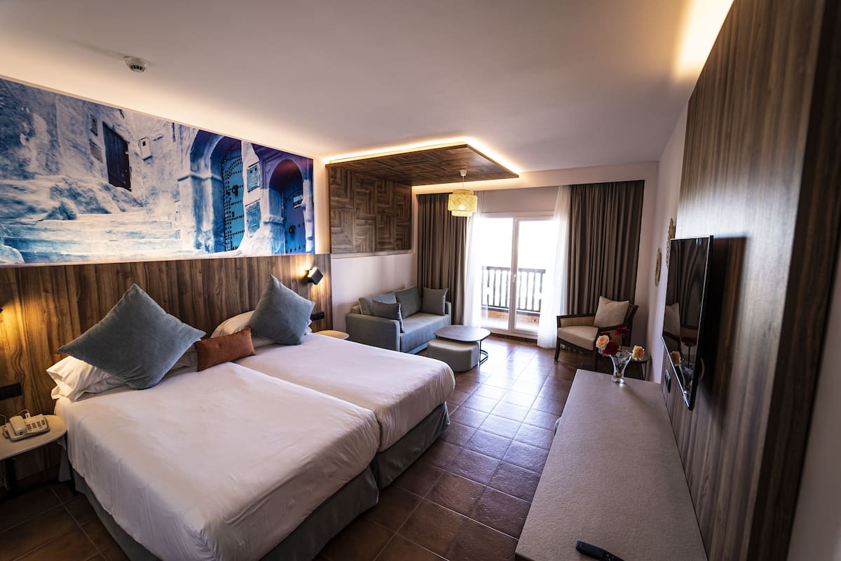 Habitación de hotel con dos camas, un sofá, una mesita, un televisor y un balcón con cortinas.