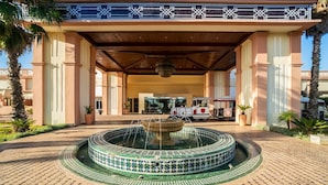 Entrada de hotel con una fuente, palmeras y un paseo techado.