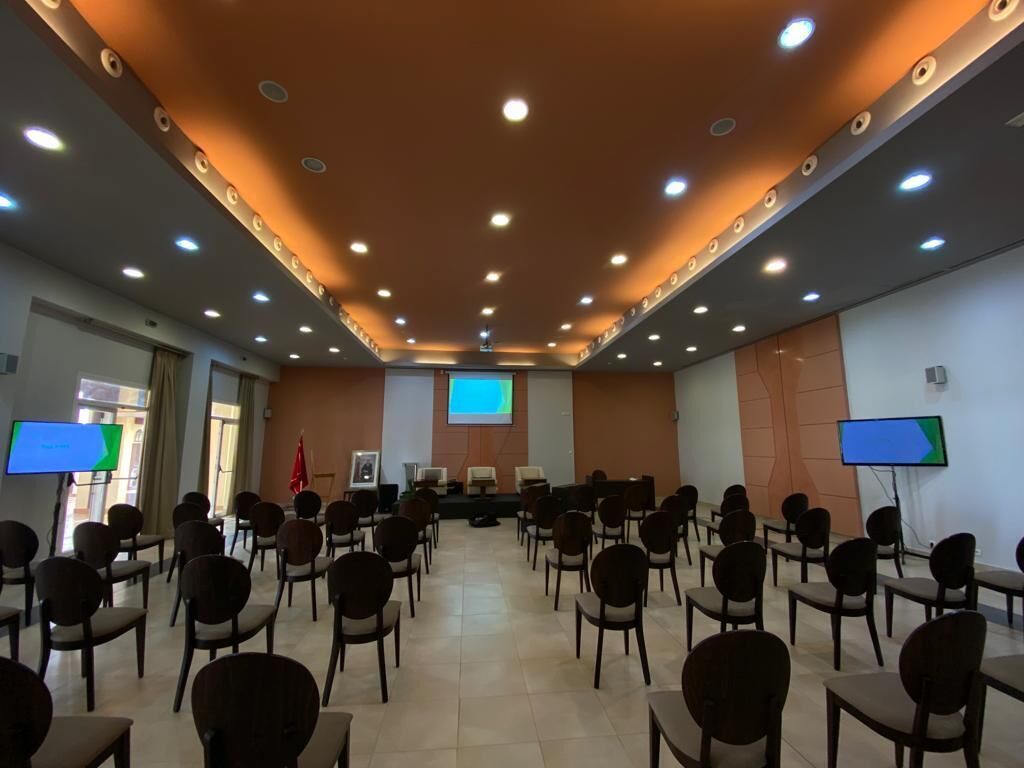 A conference room with rows of chairs, a stage with a projector, and two screens on the walls.