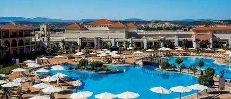 A resort with a large central pool, sun loungers, umbrellas, and a view of mountains.