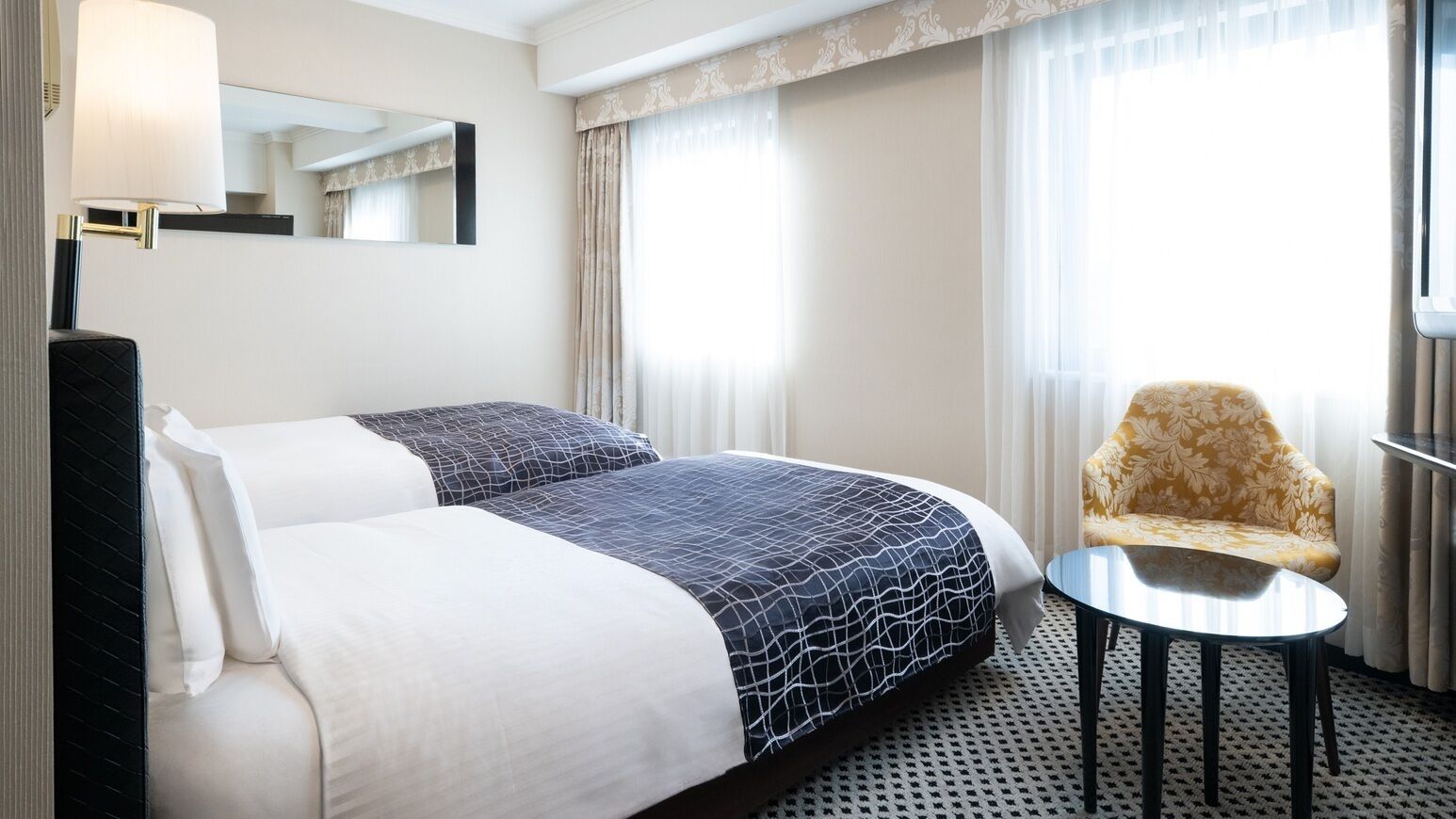 A hotel room with two beds, a chair, a small table, a lamp, and a window with curtains.