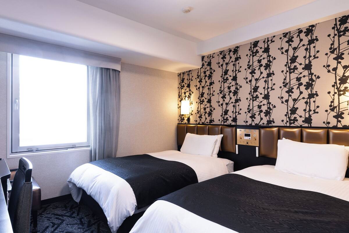 Een hotelkamer met twee bedden, een bureau, een stoel, een raam met gordijnen en een bloemenpatroon op de muur.