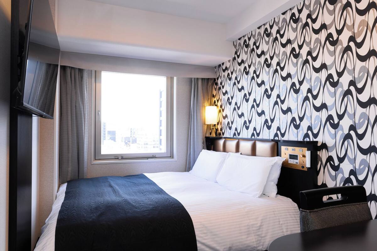 Een hotelkamer met een groot bed, een aan de muur bevestigde televisie, een raam met gordijnen en behang met een patroon.