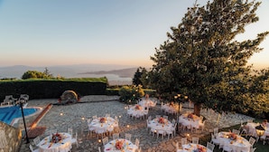 An outdoor patio with multiple round tables set up for dining, surrounded by stone flooring and greenery. - Portaria Hotel (Volos)