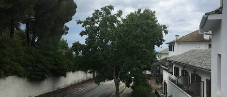 A street with a white building on the right, a tree in the center, and a parked car in the foreground.