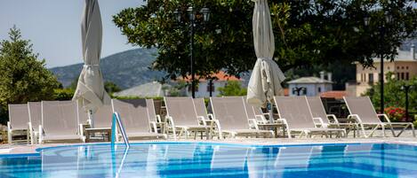 A swimming pool with lounge chairs and umbrellas, surrounded by trees and buildings in the background.