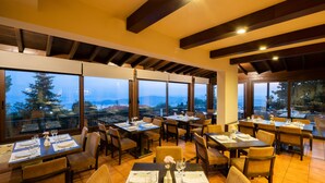 A restaurant with multiple tables set for dining, large windows offering a view of the outdoors, and a modern interior design with wooden beams on the ceiling. - Portaria Hotel (Volos)