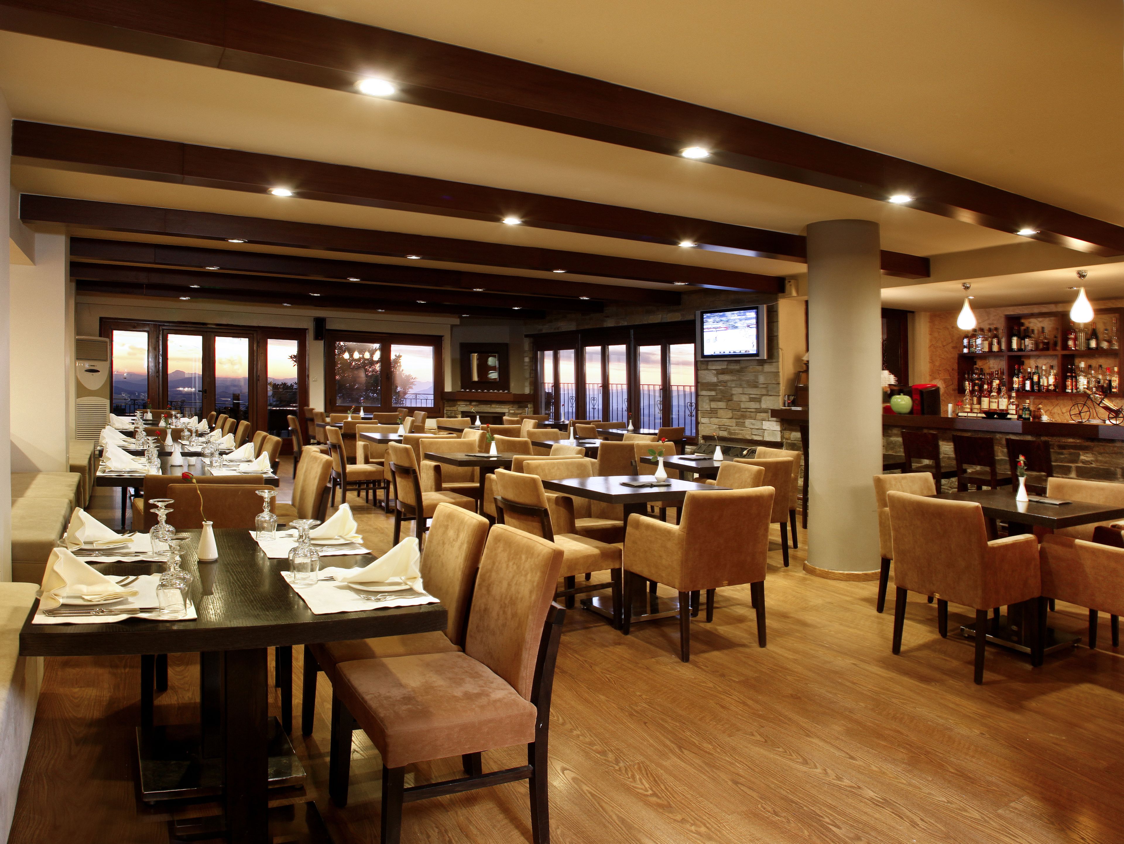 A restaurant with multiple dining tables and chairs, a bar area, and large windows offering a view.
