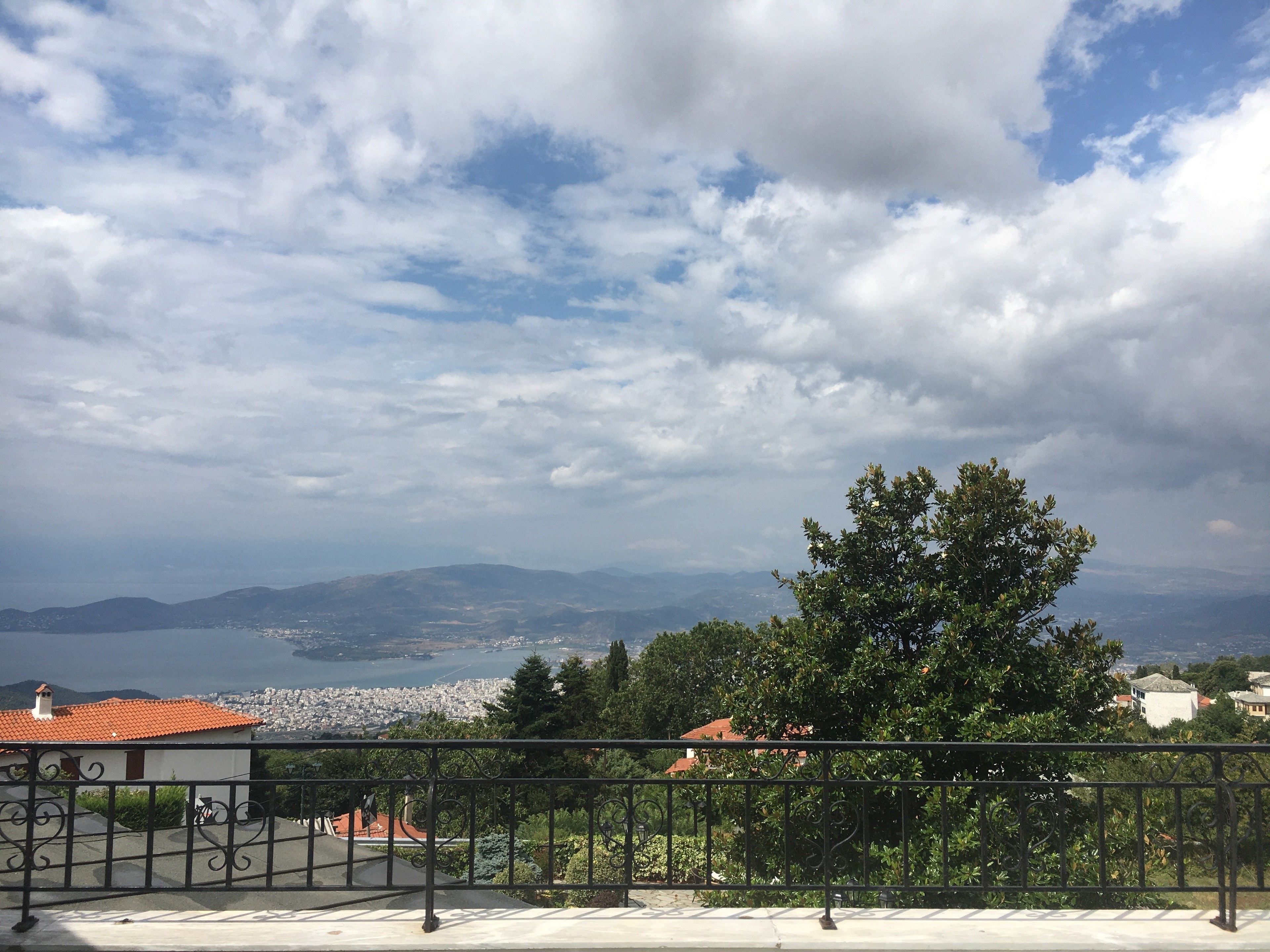 A balcony with a metal railing overlooking a cityscape, a body of water, and distant mountains.