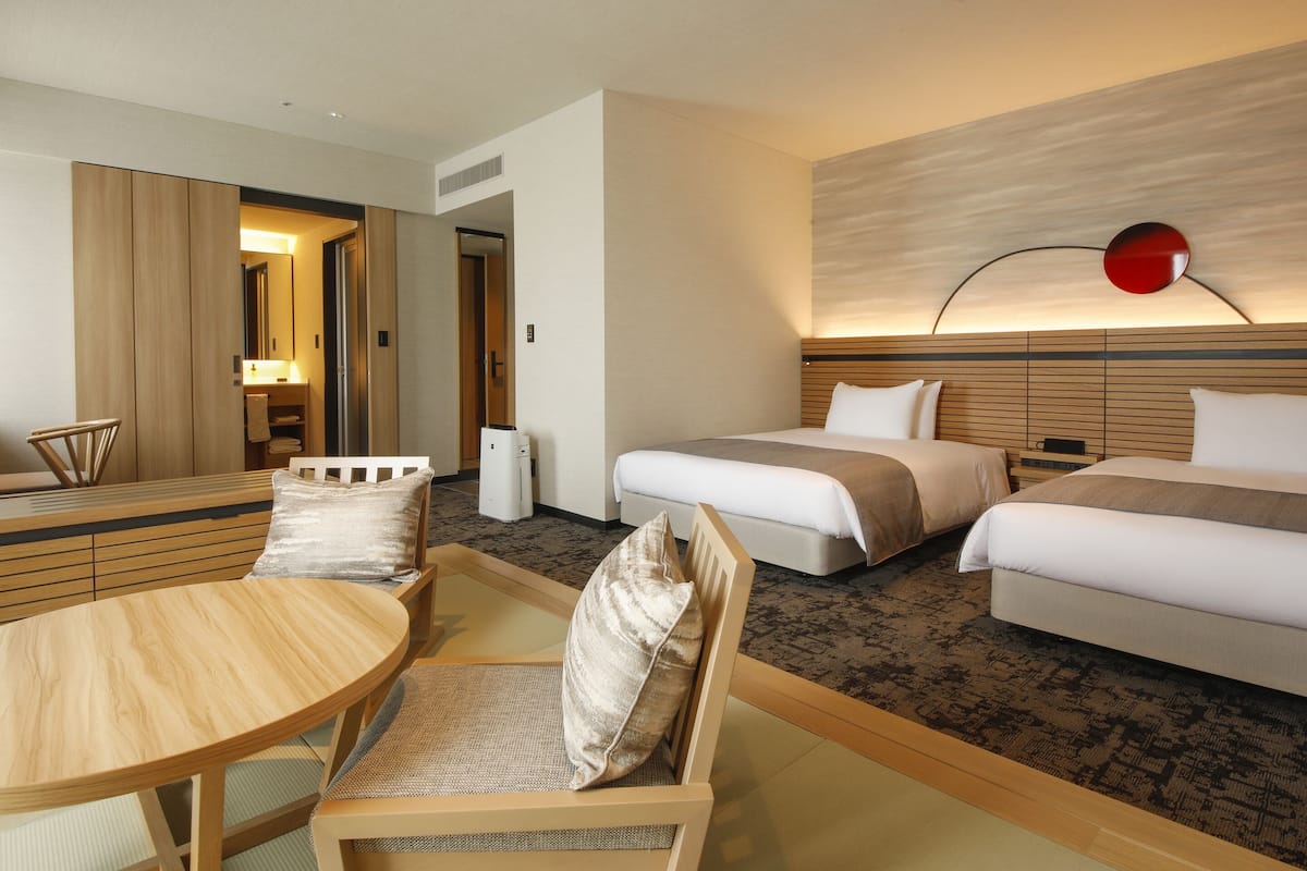 A hotel room with two beds, a wooden desk, a chair, a round table, and a modern wall lamp.