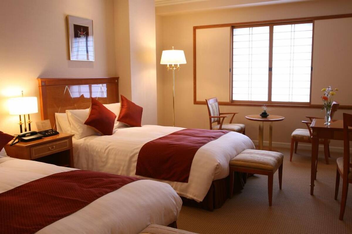A hotel room with two beds, a desk, a chair, a small table, and a window.