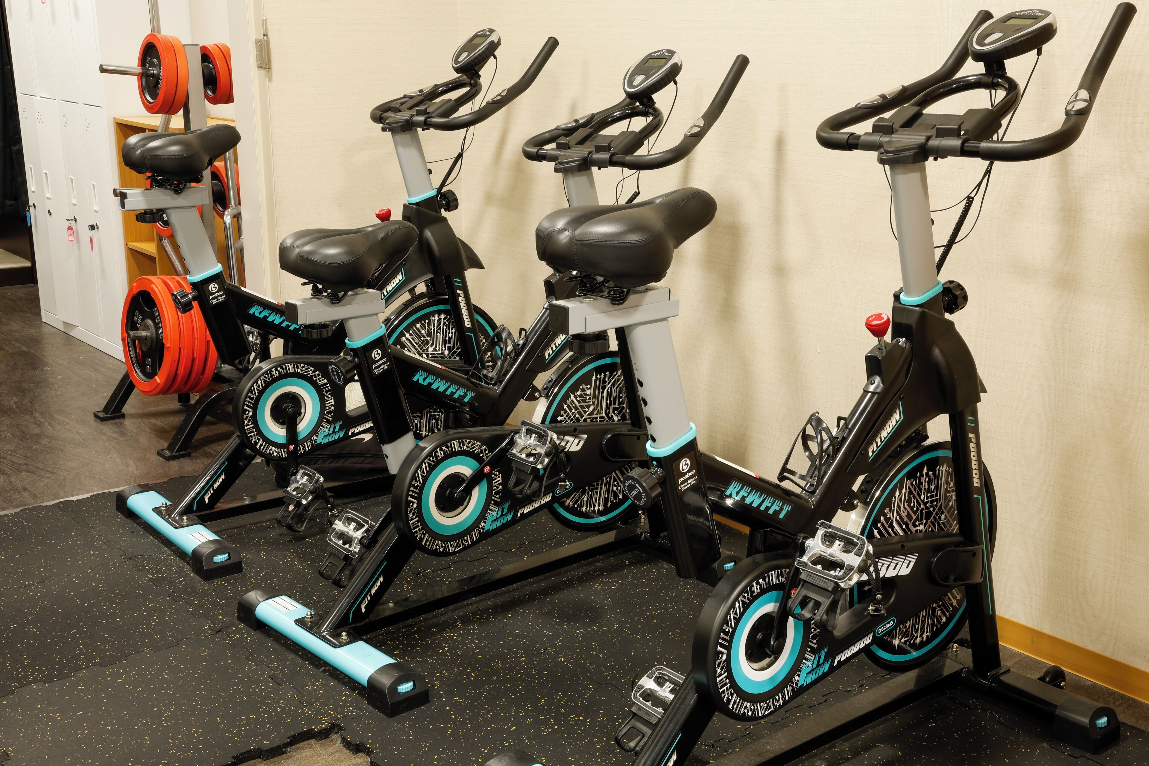 Three stationary bikes with black and teal accents, positioned in a gym with a wooden floor and a white wall.