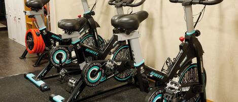 Three stationary bikes with black and teal accents, positioned in a gym with a wooden floor and a white wall.