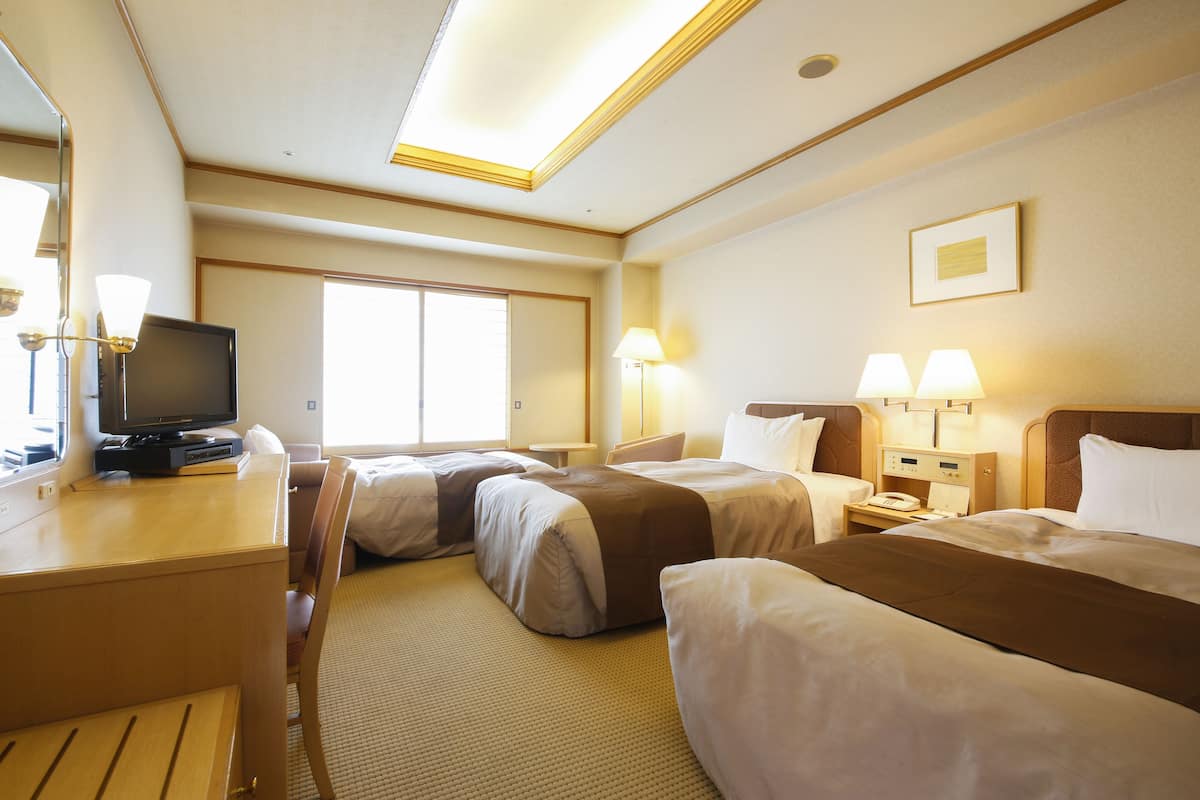 A hotel room with two beds, a desk with a television, bedside lamps, and a large window.