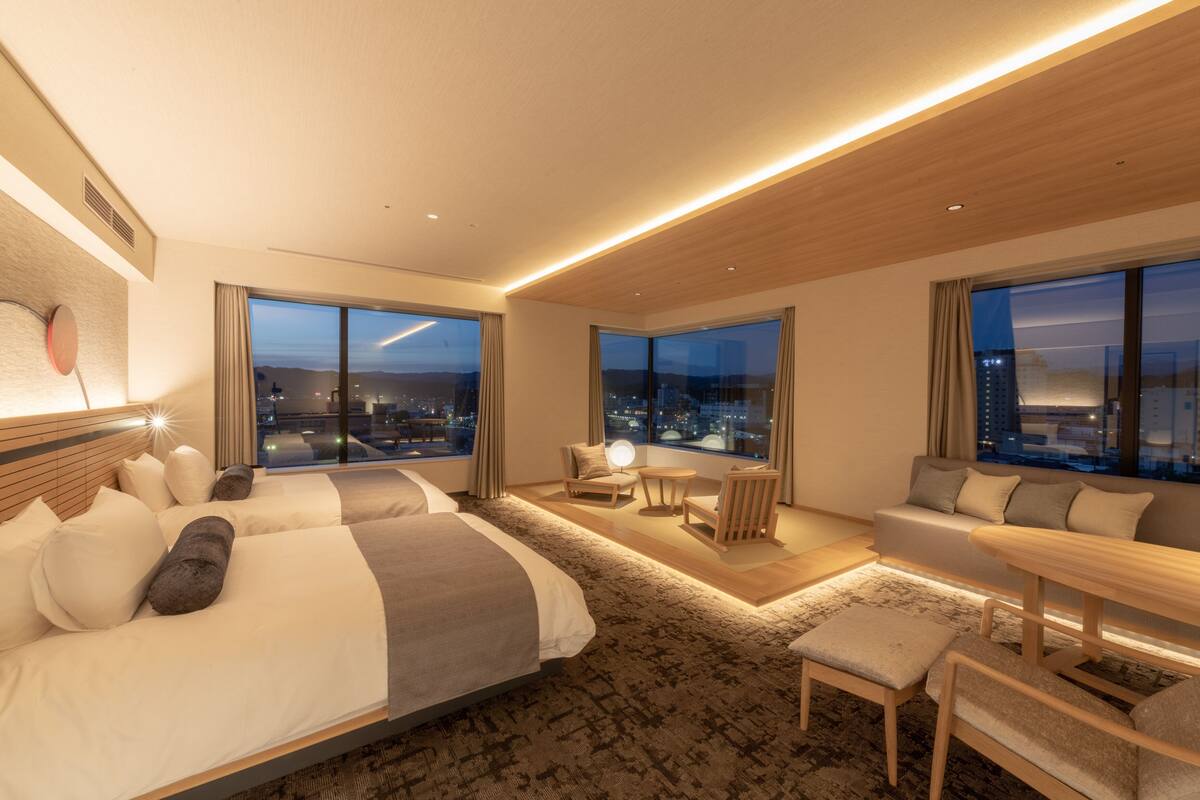 A modern hotel room with a large bed, a seating area with a table, and large windows offering a city view.