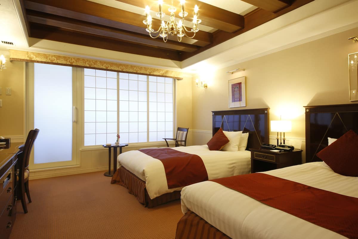 A hotel room with two beds, a desk, a chair, a window with blinds, a chandelier, and a nightstand with a lamp.
