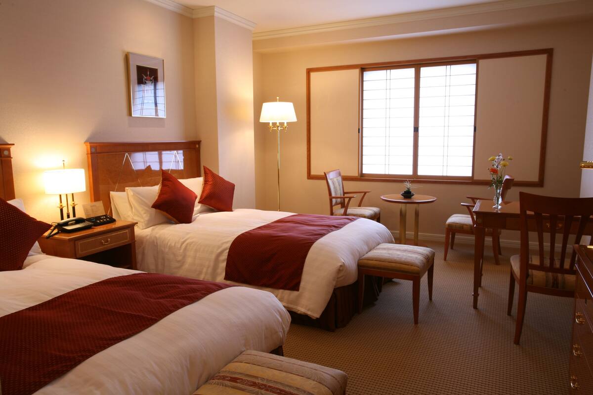 A hotel room with two beds, a desk, a chair, a lamp, and a window.