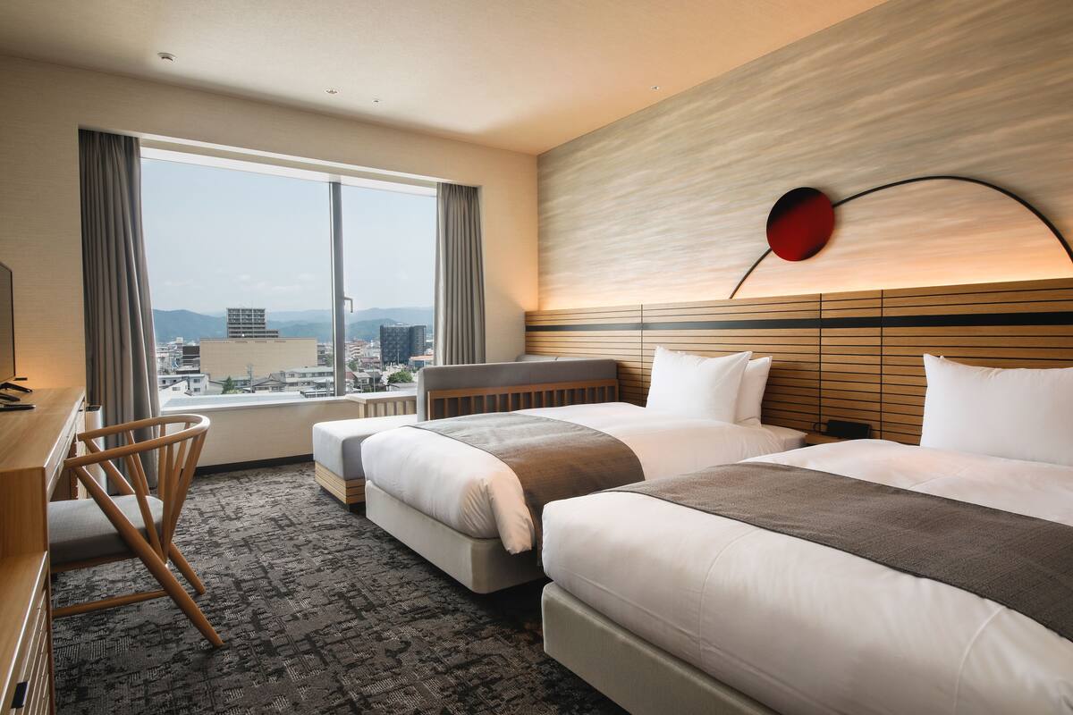A hotel room with two beds, a desk, and a chair. The room has a large window with a city view.