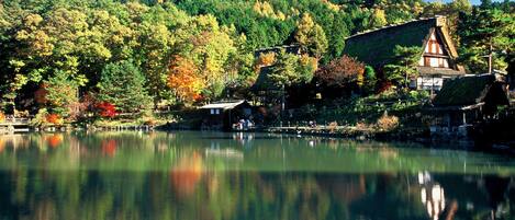 A traditional Japanese house with a sloped roof, located beside a calm lake with reflections of trees and buildings.