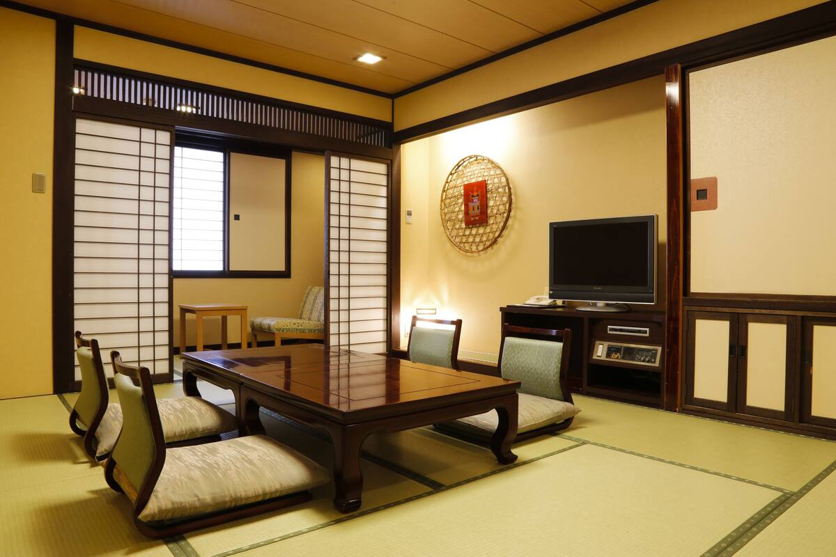 A traditional Japanese room with tatami flooring, a low table with cushions, a television, and a decorative wall hanging.